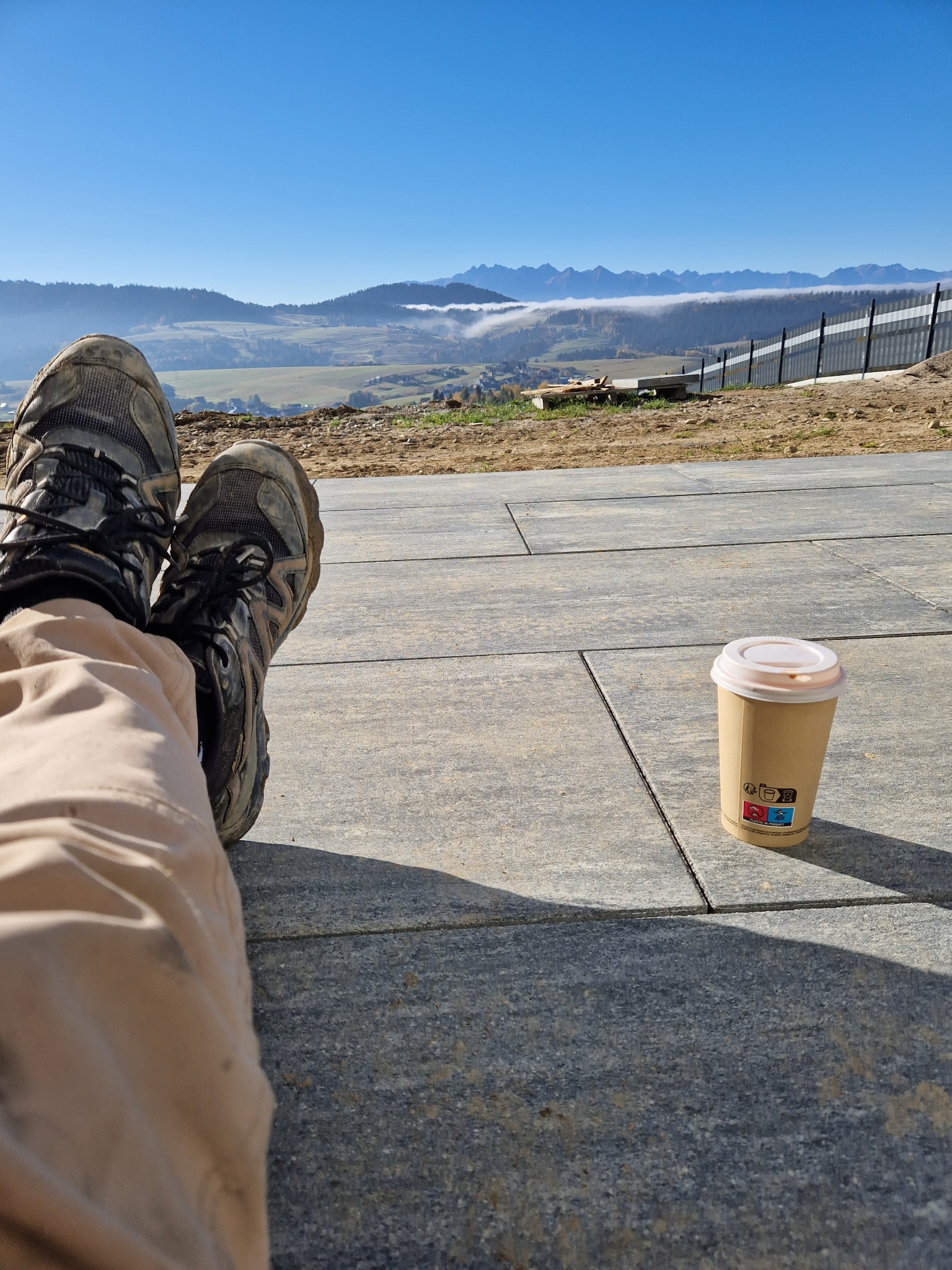 Przerwa na kawę na ułożonej szarej kostce brukowej z widokiem na góry w oddali, wystające nogi w brudnych butach roboczych.