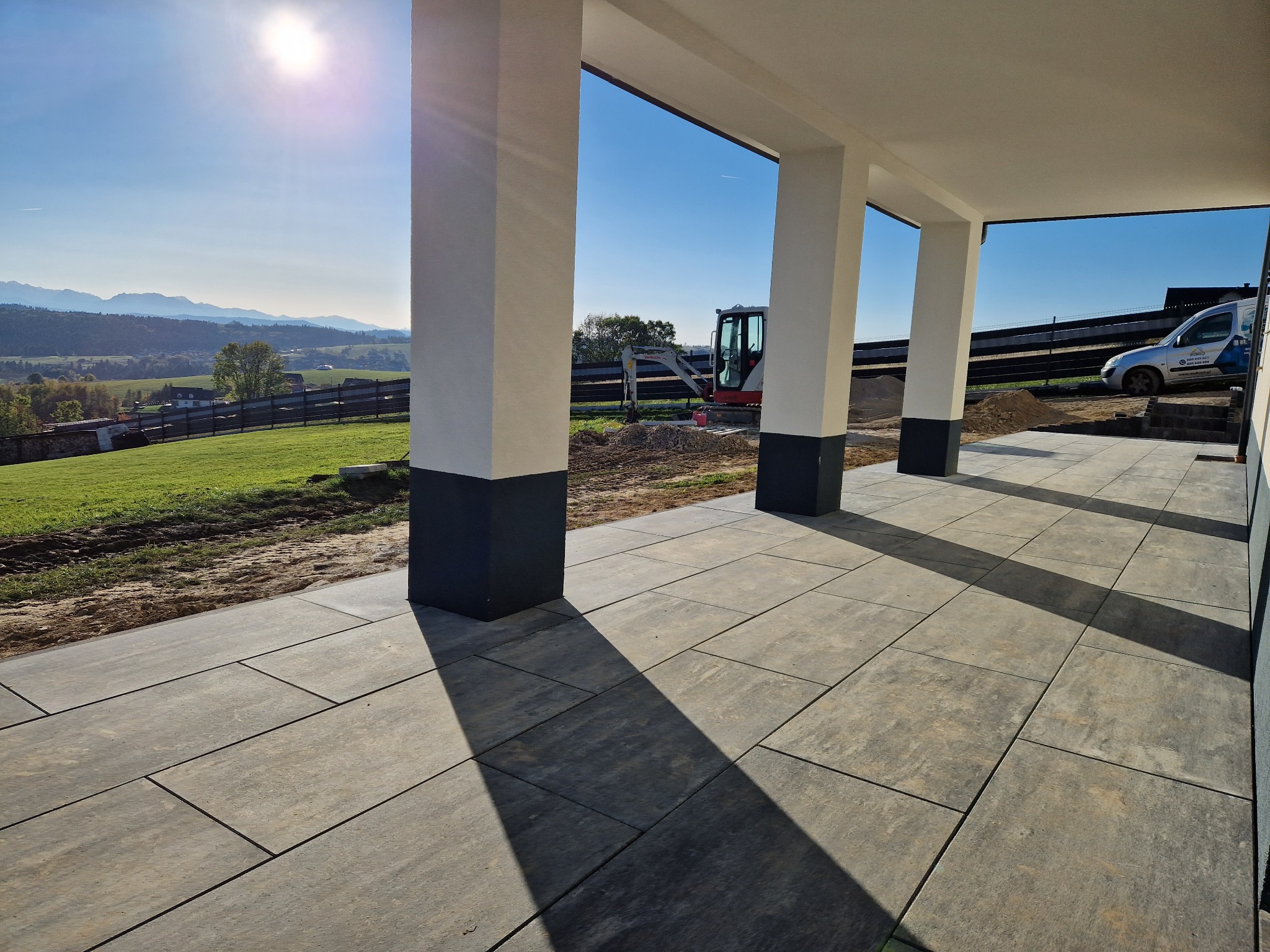 Ułożona kostka brukowa w odcieniach szarości na tarasie z widokiem na krajobraz górski i prace ziemne w tle, oświetlona porannym słońcem.