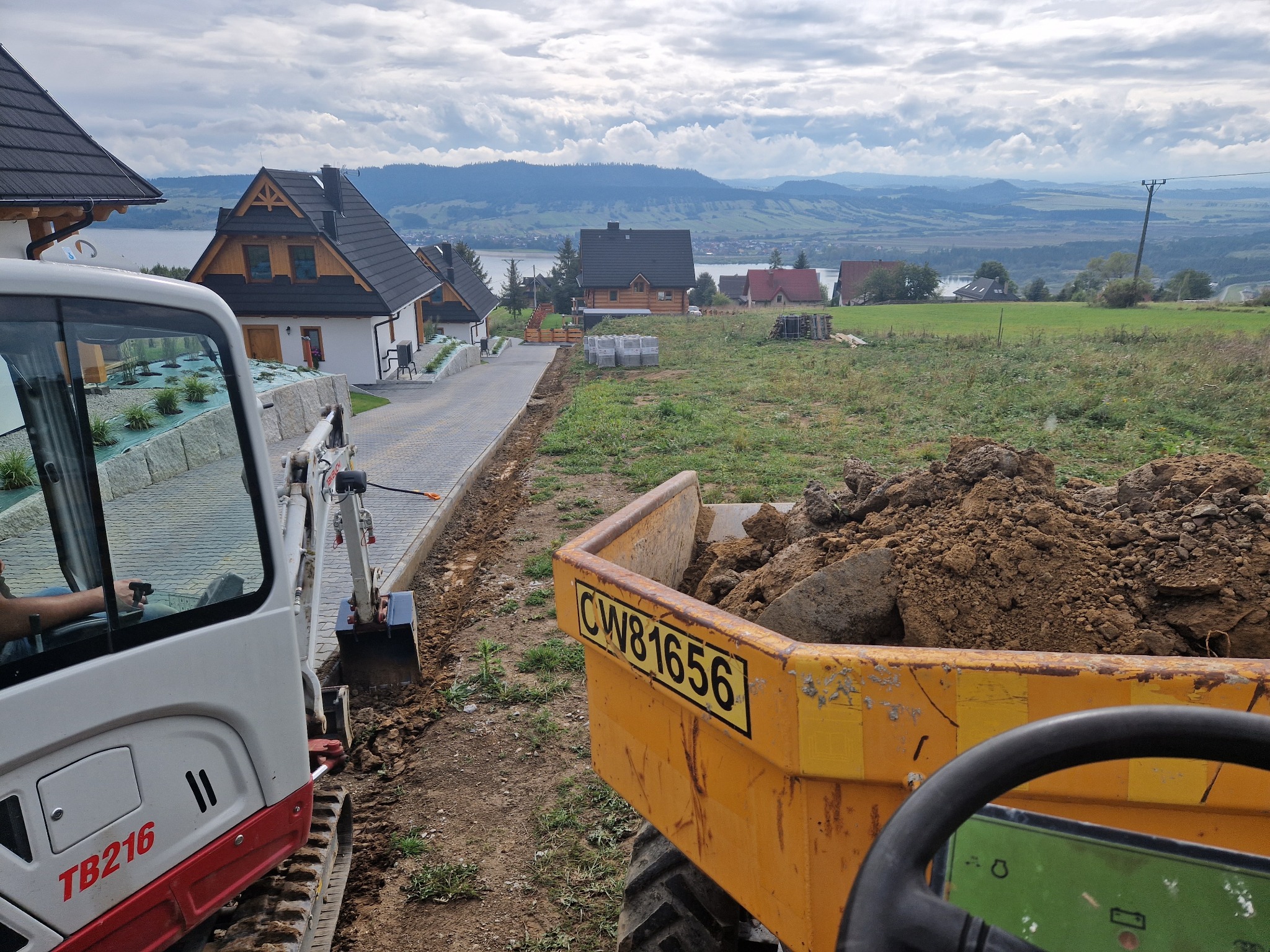 Mini koparka TB216 i wywrotka CWB1656 w trakcie prac ziemnych przy budowie drogi dojazdowej z kostki w malowniczym krajobrazie górskim.