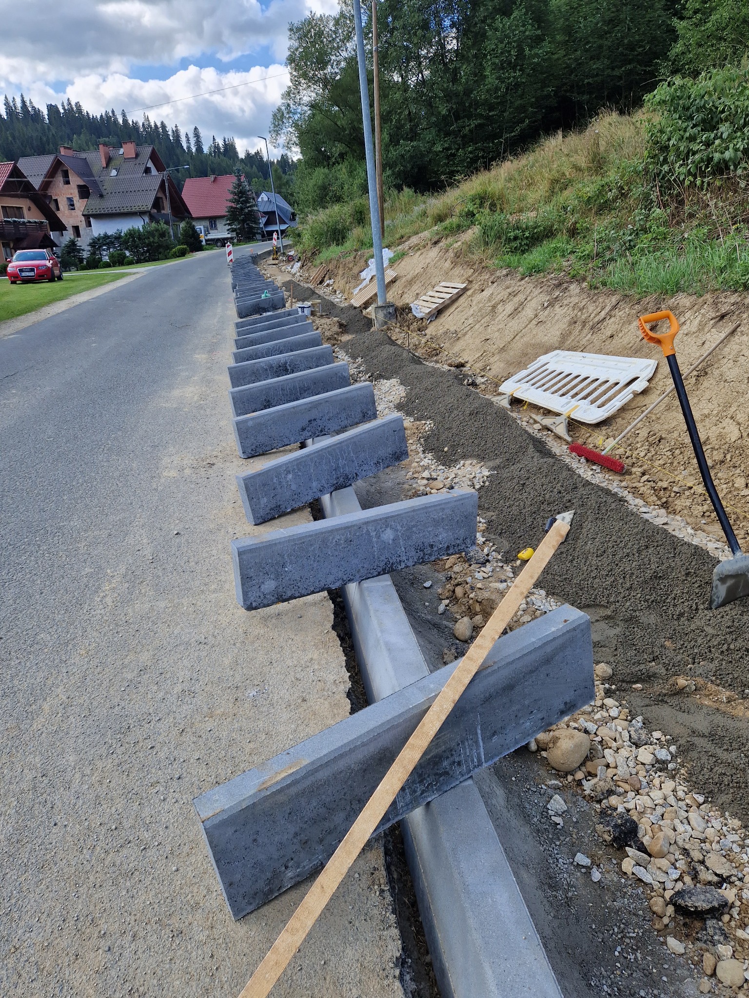 Montaż betonowych obrzeży chodnika przy asfaltowej drodze, widoczne narzędzia i materiały budowlane, w tle domy i zalesione wzgórze.
