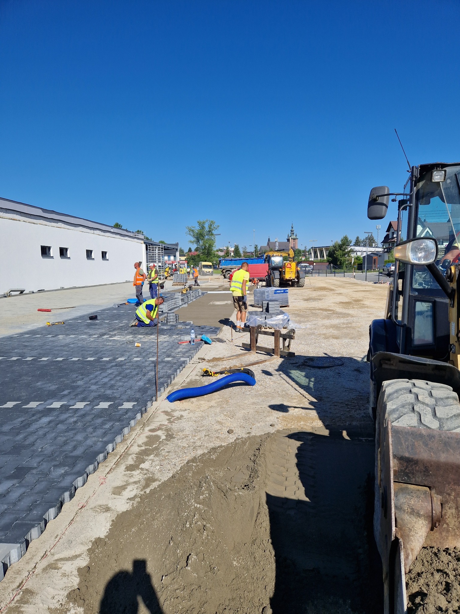 Pracownicy w odblaskowych kamizelkach układają szarą kostkę brukową na przygotowanym podłożu; w tle budynek, niebo bezchmurne, oraz ciężki sprzęt budowlany