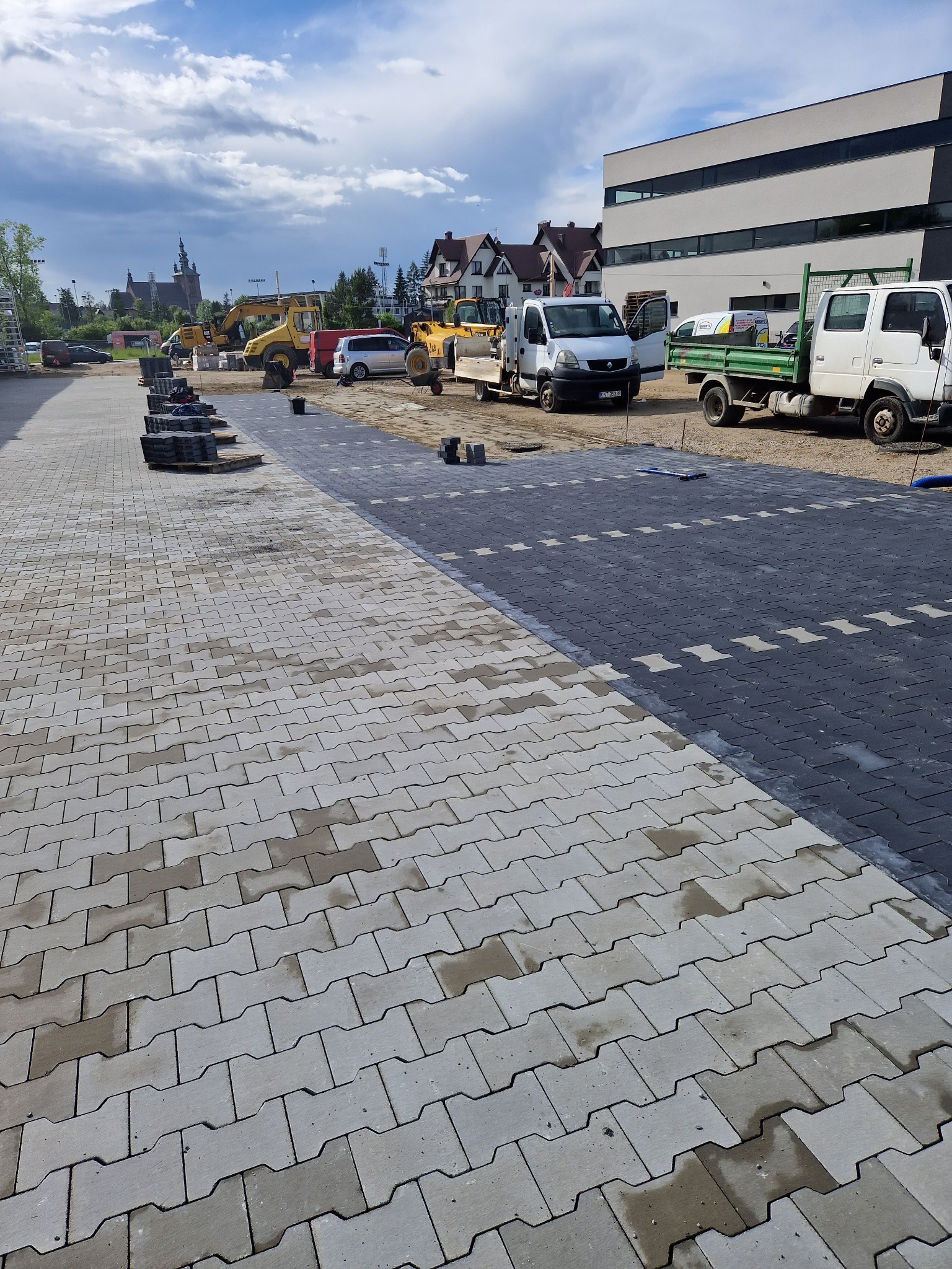 Częściowo ułożona szara kostka brukowa typu 'I' na placu budowy, widoczne maszyny budowlane i zaparkowane samochody dostawcze, w tle budynek z szarymi pasami i kościół.