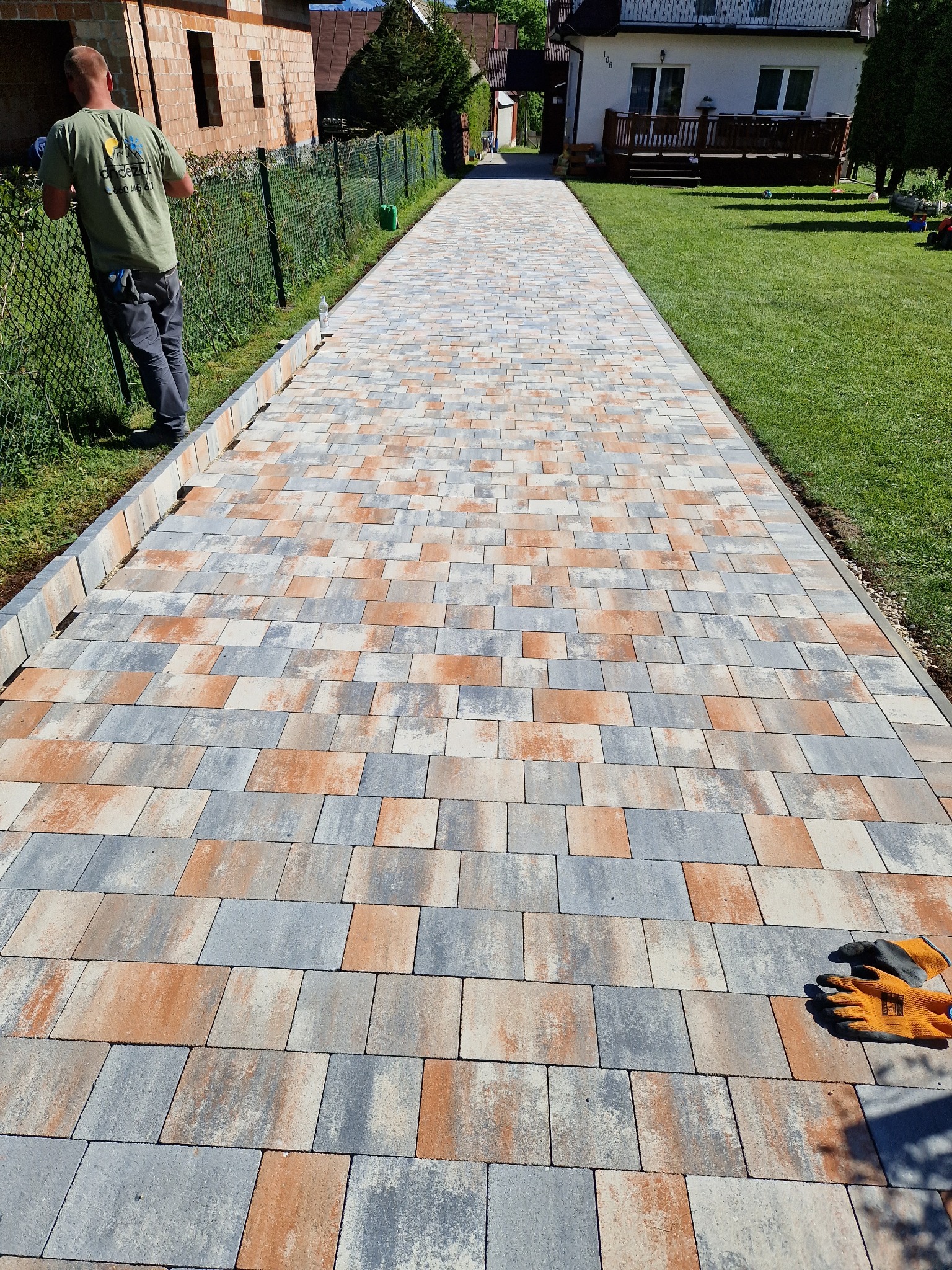 Długa, nowo ułożona ścieżka z kostki brukowej w odcieniach szarości i pomarańczy prowadząca przez trawnik w kierunku domu, widoczne rękawice robocze na kostce i pracownik przy ogrodzeniu.