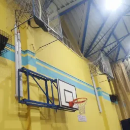 Wewnętrzna instalacja oświetleniowa hali sportowej z lampami zabezpieczonymi metalowymi koszami, widoczne również rury i fragment konstrukcji kosza do koszykówki.