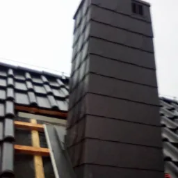 Nowy komin obłożony ciemną blachą, widok z dołu na tle szarego nieba, fragment dachu z dachówką w tle, widoczne elementy konstrukcji drewnianej przy kominie.