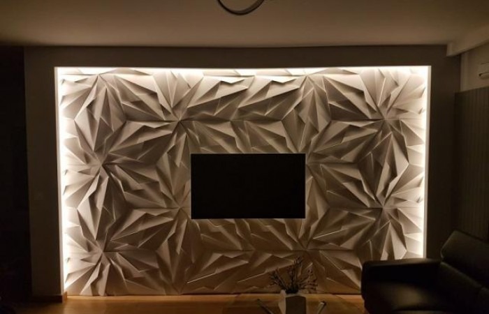 Ściana dekoracyjna z trójwymiarowych paneli o geometrycznym wzorze, podświetlona listwą LED, z miejscem na telewizor w centrum. Widoczny fragment sofy i stolika kawowego.
