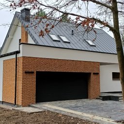 Erbud Robert Strus - Nowoczesny dom z garażem, wykończony cegłą klinkierową i białym tynkiem. Dach z blachy z dwoma oknami dachowymi. Brukowany podjazd i zagospodarowany teren.