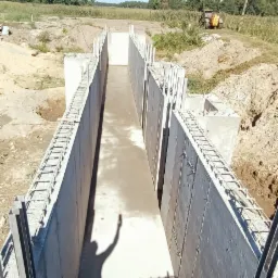 Świeżo wylany beton w szalunku fundamentowym, z widocznym zbrojeniem i cieniem osoby wznoszącej rękę, na tle krajobrazu z traktorem i drzewami.