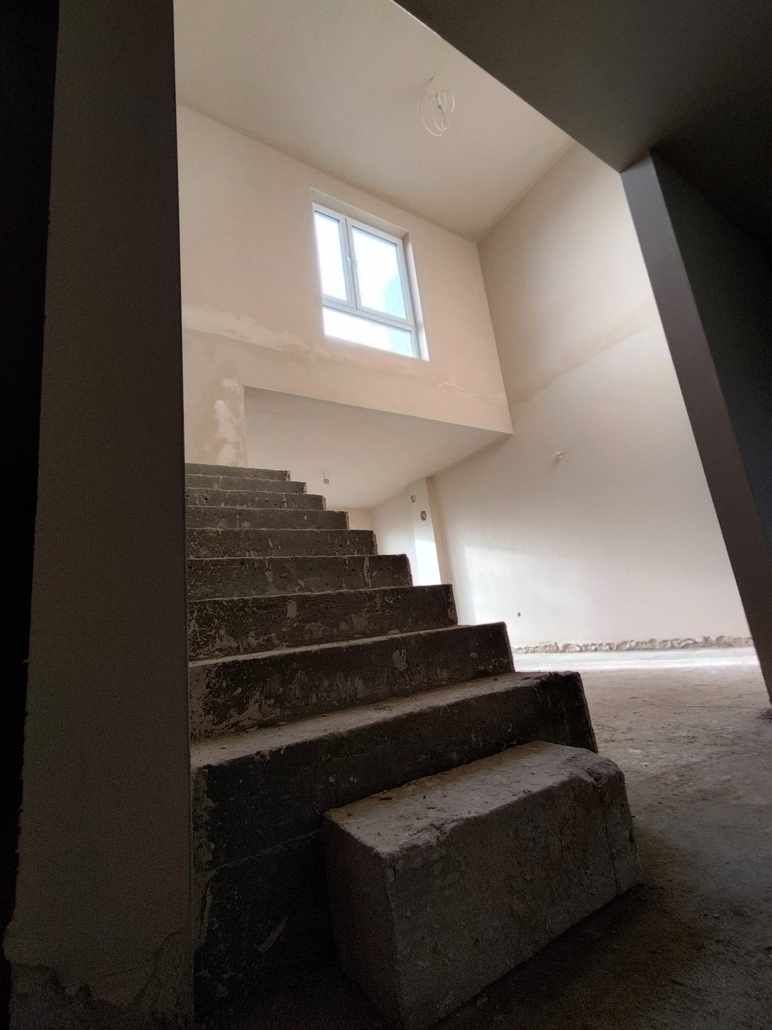 Wnętrze w trakcie prac wykończeniowych: surowe schody betonowe prowadzą na górę, widoczne okno i przygotowane do malowania ściany. Stan deweloperski.