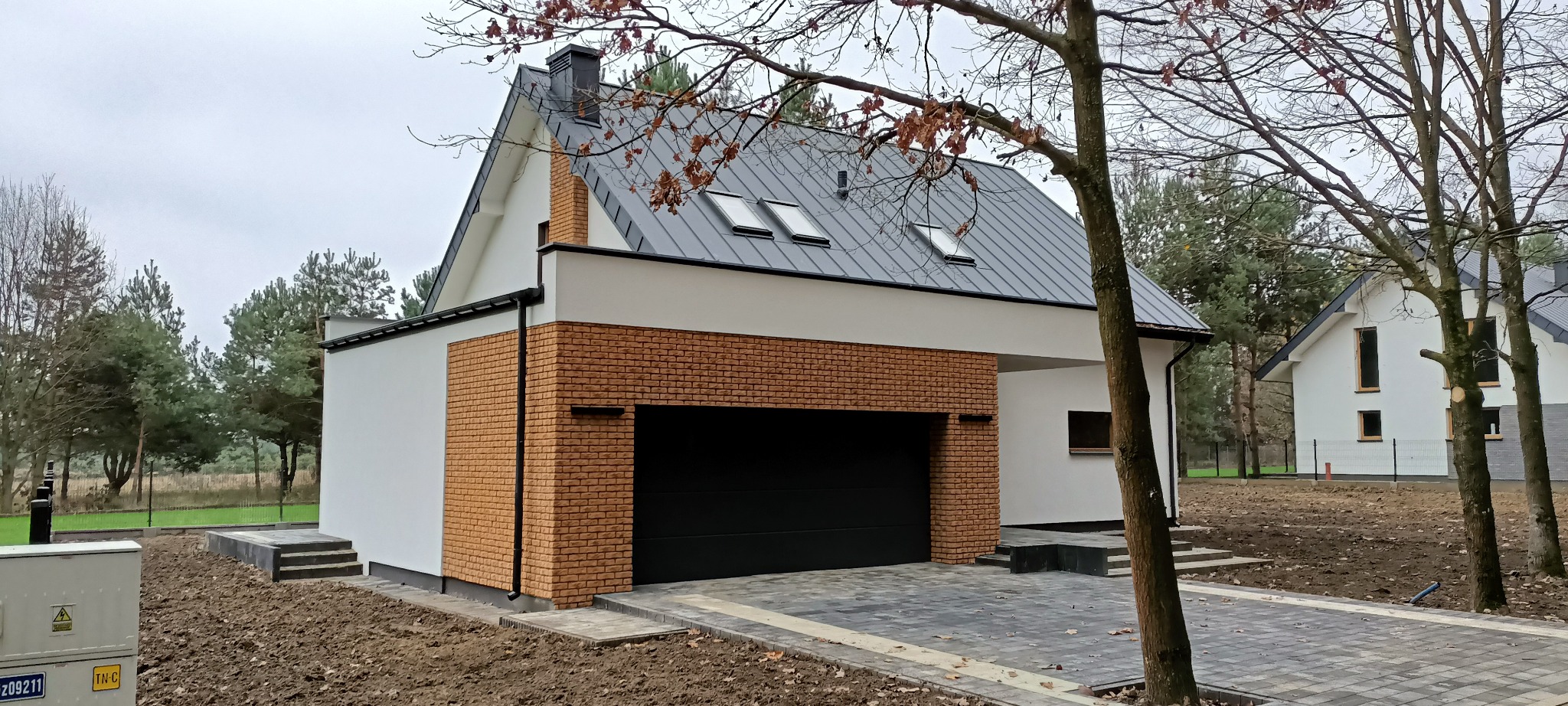 Nowoczesny dom z garażem, wykończony cegłą klinkierową i białym tynkiem. Dach z blachy z dwoma oknami dachowymi. Brukowany podjazd i zagospodarowany teren.