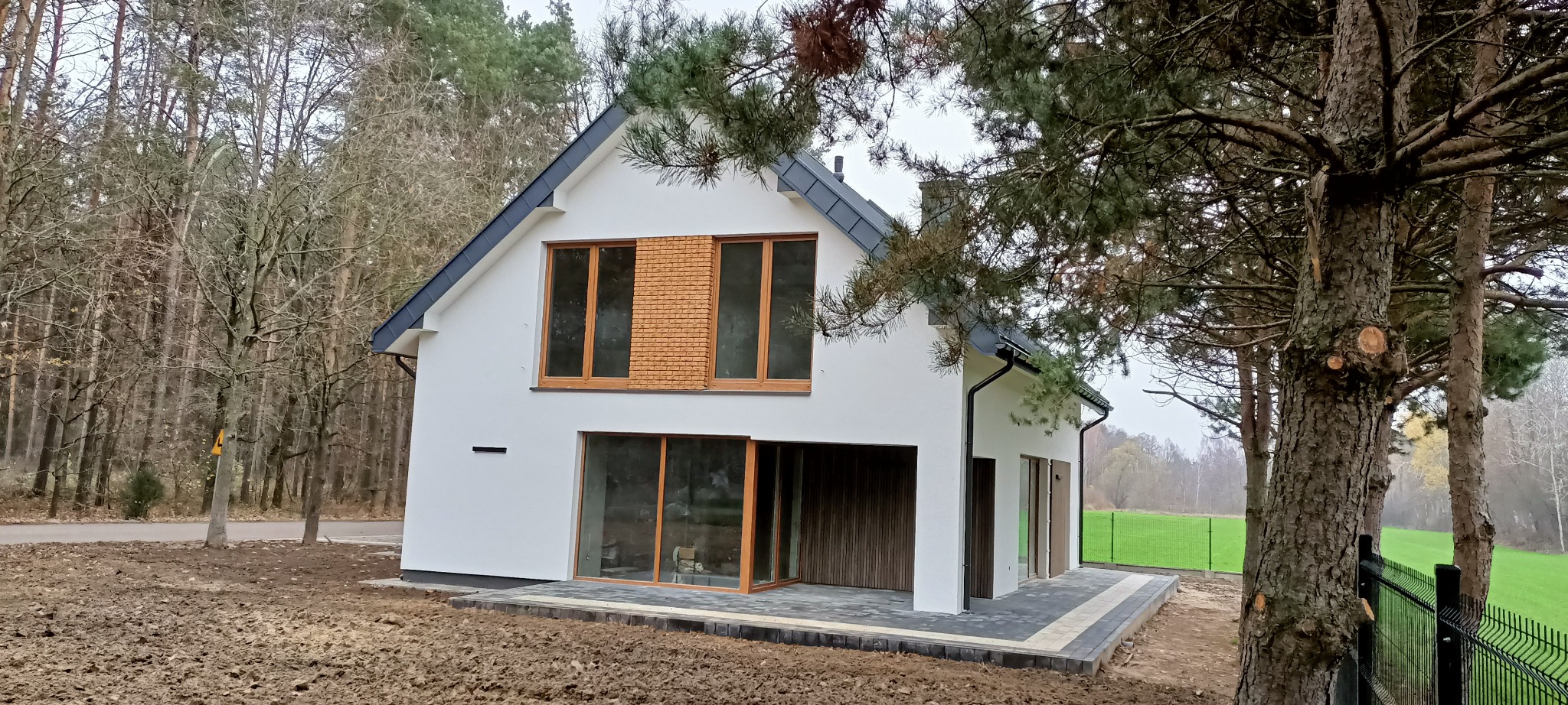 Nowoczesny dom z białymi ścianami, drewnianymi oknami i szarą dachówką, otoczony zielenią i drzewami. Widoczne detale wykończenia elewacji i zagospodarowania terenu.