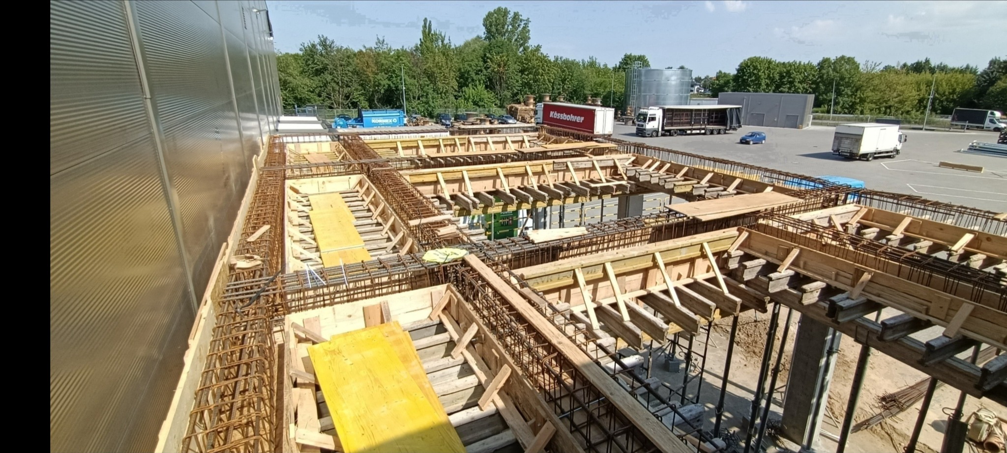 Zbrojenie fundamentów pod budynek przemysłowy z widocznymi deskowaniami i stalowymi prętami, w tle zaparkowane ciężarówki i budynek magazynowy.