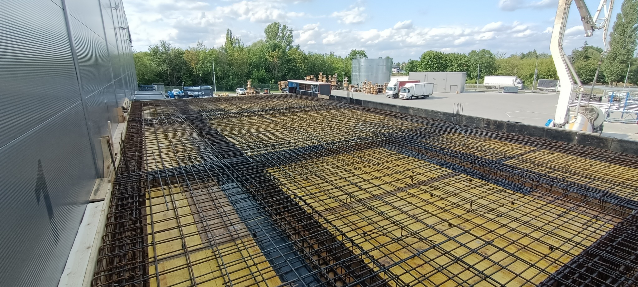 Siatka zbrojeniowa na deskowaniu fundamentu budynku przemysłowego, widoczna z góry, z budynkiem w tle i ciężarówką z pompą do betonu.
