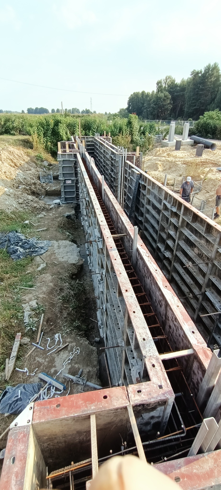 Szalunki zbrojeniowe przygotowane do zalania betonem, widok z góry na wykop, w tle zieleń i dwóch pracowników budowlanych.