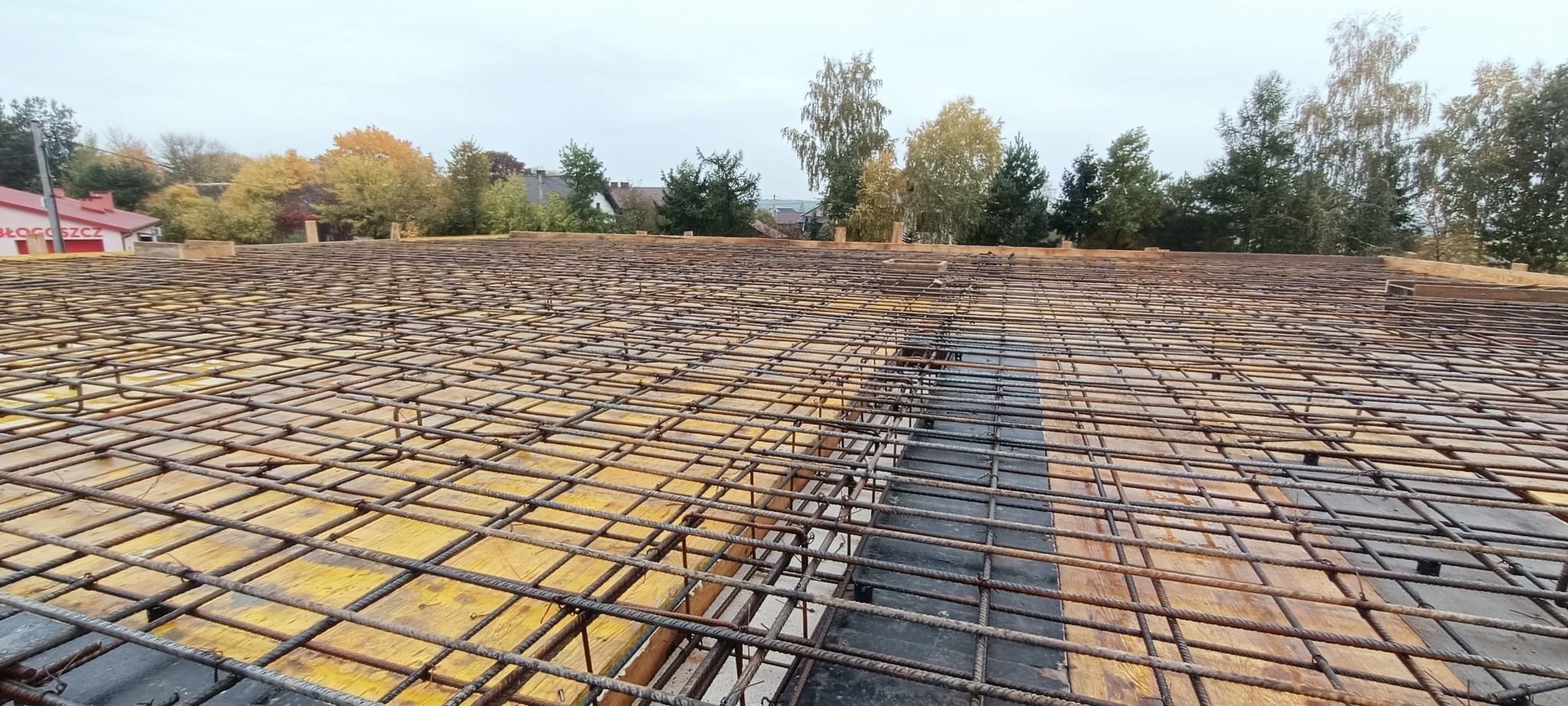 Zbrojenie płyty fundamentowej z widoczną siatką stalową na deskowaniu, przygotowane do zalania betonem, z widokiem na zabudowania i drzewa w tle.