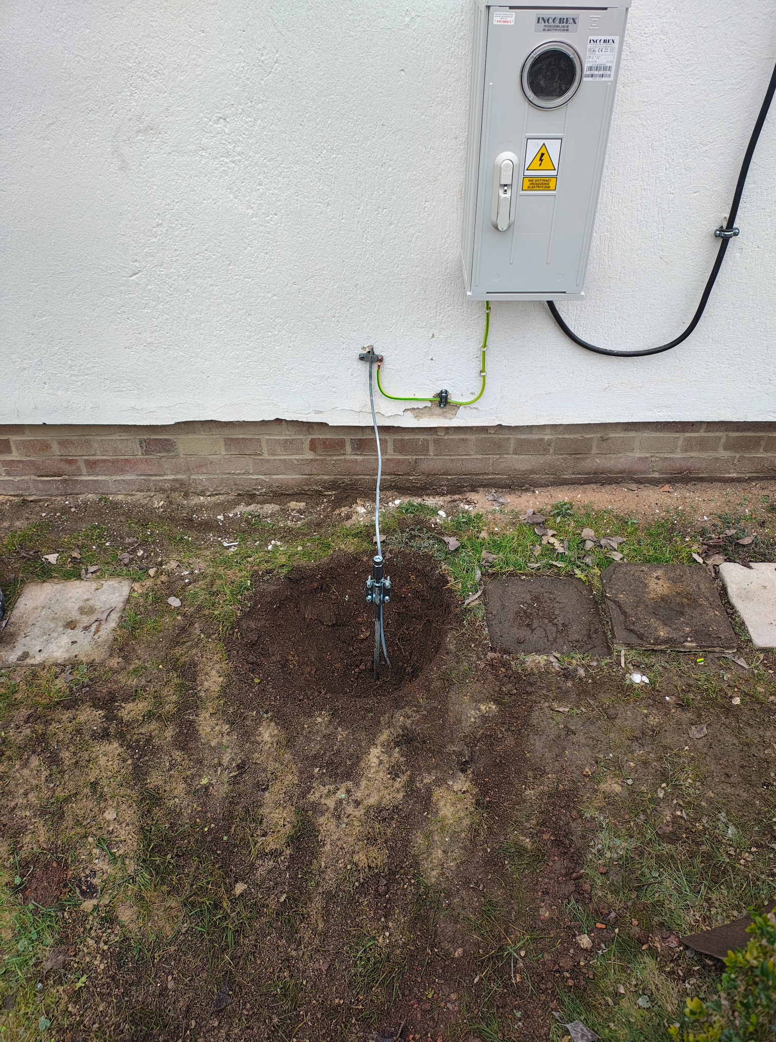Uziemienie budynku z szarą skrzynką elektryczną na białej ścianie. Widoczny kabel uziemiający i wbity w ziemię element uziemiający. Prace ziemne wokół uziemienia.