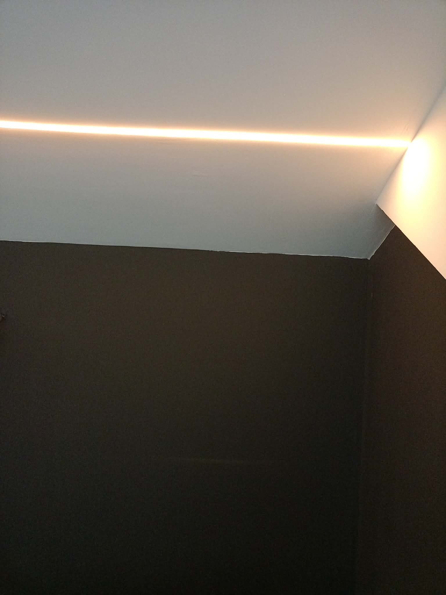 Minimalistyczne oświetlenie LED wzdłuż sufitu, ciepłe światło podkreśla krawędź ściany. Nowoczesny design wnętrza z ciemną ścianą i jasnym sufitem.