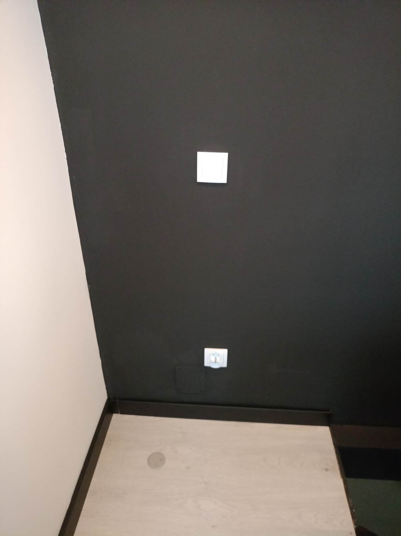 Czarne ściany z dwoma białymi gniazdkami elektrycznymi, widoczne także fragmenty białej ściany i podłogi z listwą przypodłogową. Minimalistyczne wnętrze.