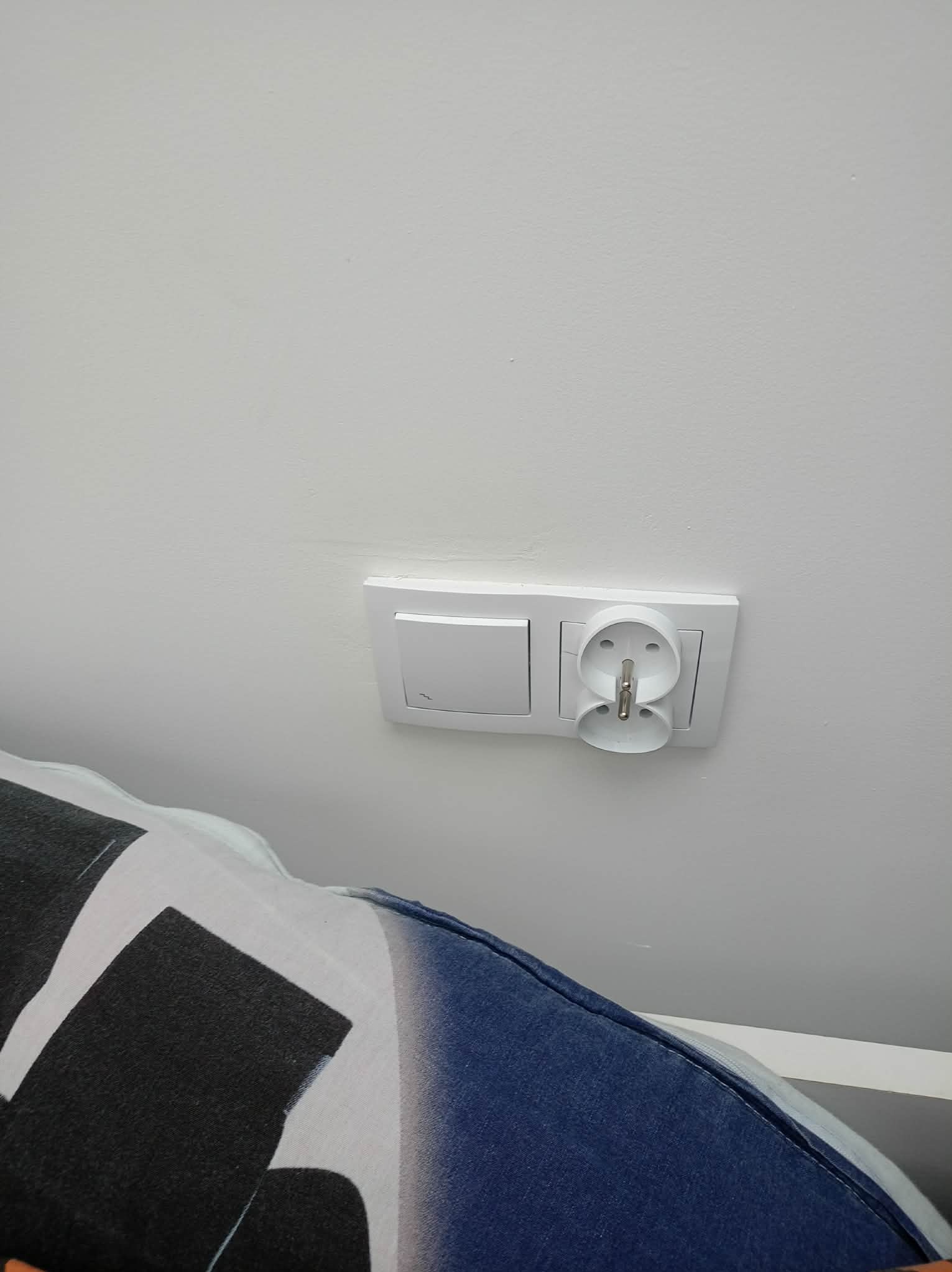 Biały, podwójny kontakt elektryczny z włącznikiem światła na jasnej ścianie. Widoczny fragment ubrania osoby robiącej zdjęcie.
