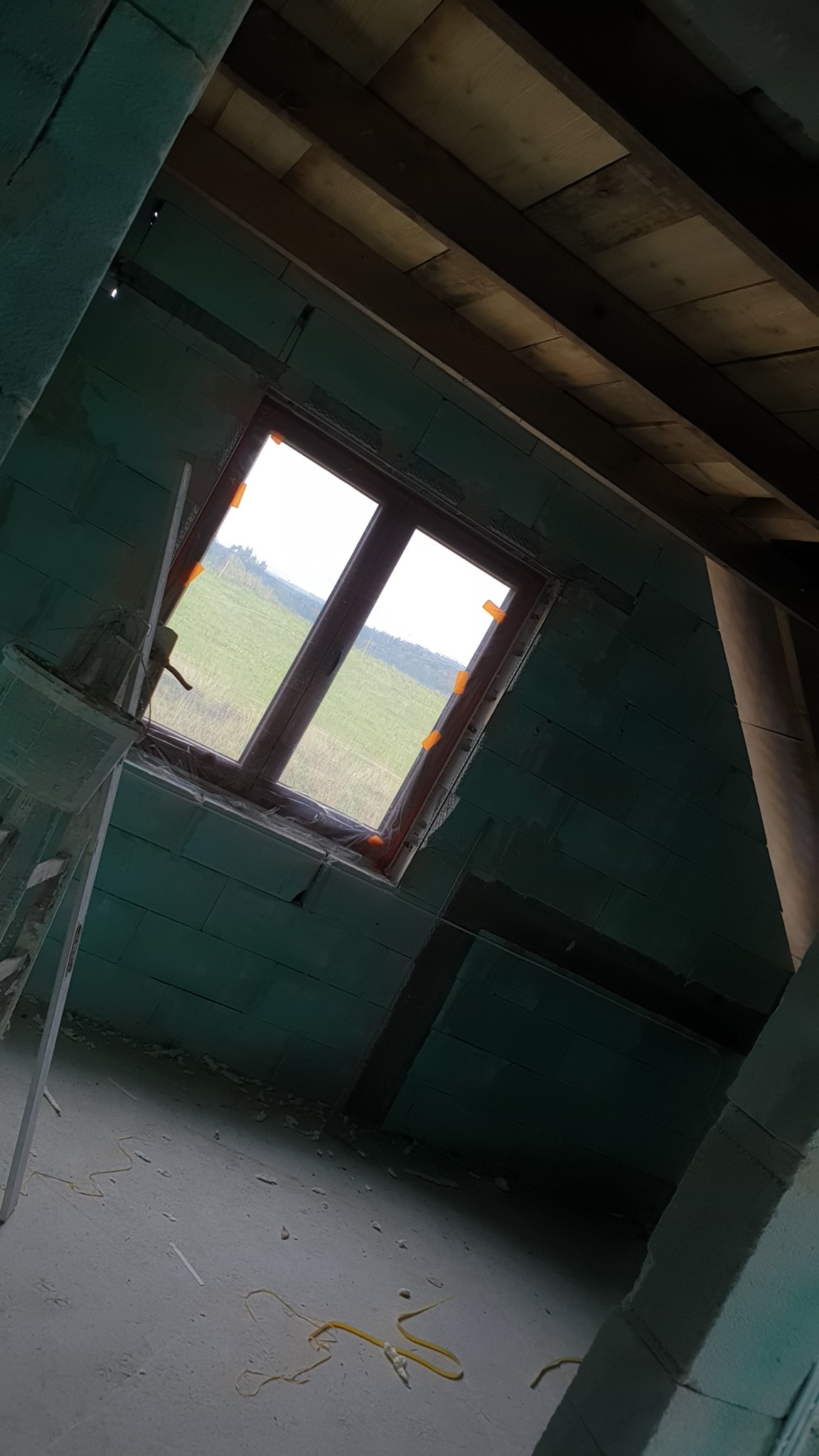 Surowy stan wnętrza z oknem wychodzącym na zielone pole, ściany z pustaków, widoczne drewniane belki stropowe, podłoga w stanie roboczym z resztkami materiałów budowlanych, okno zabezpieczone folią.