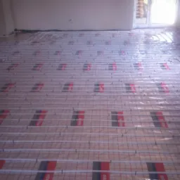 Białe rurki ogrzewania podłogowego ułożone na folii z czerwono-czarnymi oznaczeniami, widoczne z perspektywy wejścia do pomieszczenia, z oknem w tle.