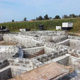 Wykopane fundamenty domu jednorodzinnego z ułożonymi bloczkami betonowymi i zbrojeniami stalowymi, widok z góry na tle pola kukurydzy.