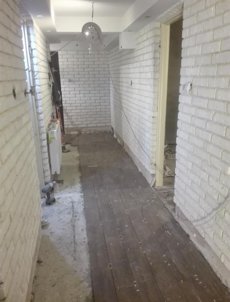 Wąski korytarz w trakcie remontu, z fragmentem ułożonej ciemnobrązowej podłogi z płytek imitujących drewno, białe ściany wykończone cegłą, widoczny wiertarko-wkrętak przy ścianie, brak listew...