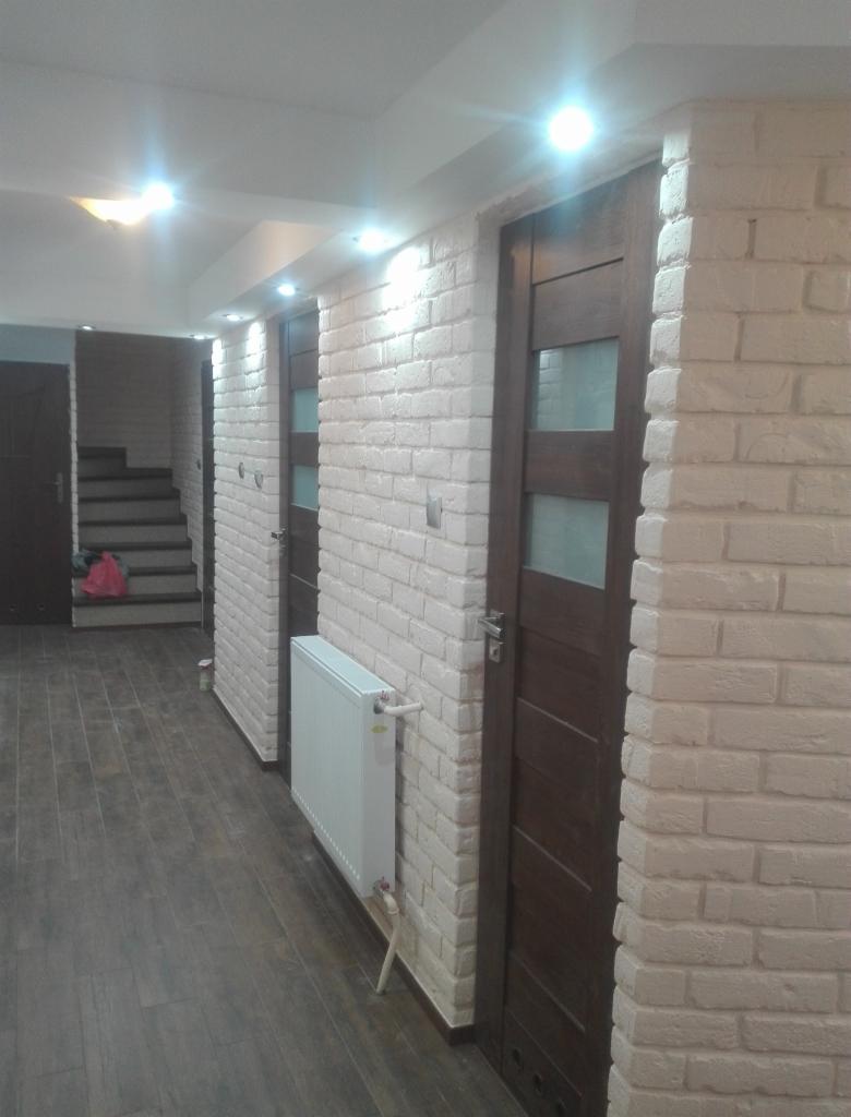 Korytarz po renowacji z białymi ścianami z cegły, ciemnymi panelami podłogowymi i drzwiami z przeszkleniami. Widoczny grzejnik i schody w oddali.