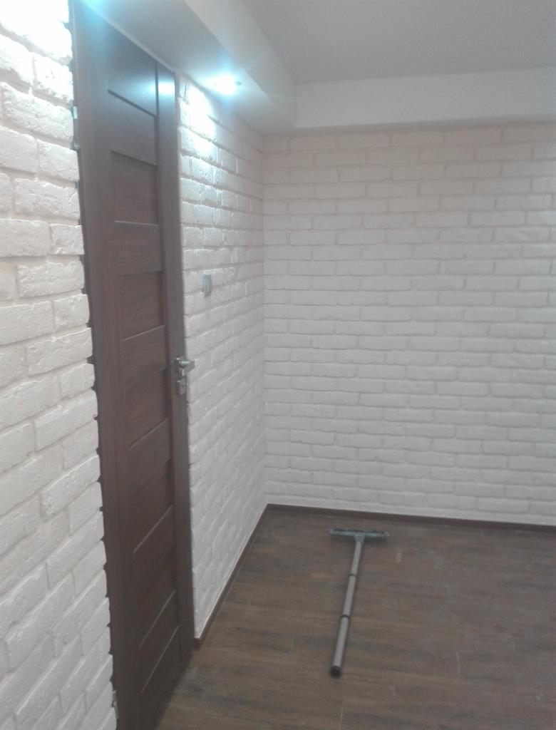 Wnętrze pomieszczenia z białą cegłą na ścianach, brązowymi panelami podłogowymi i ciemnymi drzwiami; widoczny mop do podłogi.