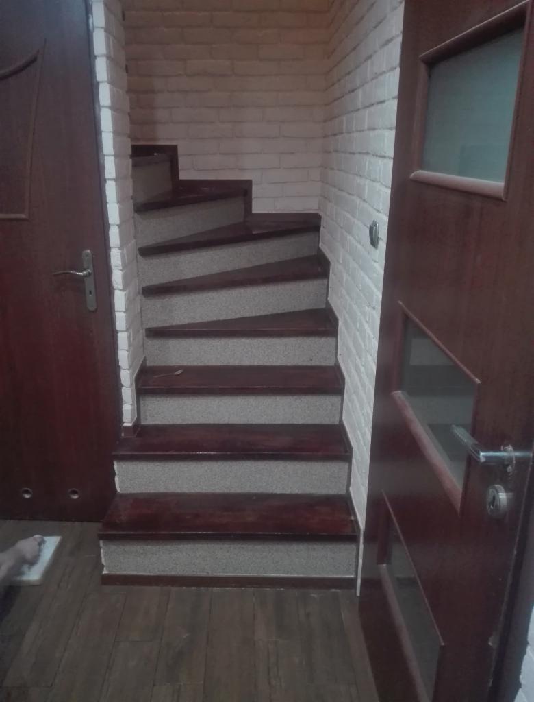 Klatka schodowa z drewnianymi stopniami i szarymi podstopnicami, białe ściany z cegły, widoczne dwie drewniane drzwi.