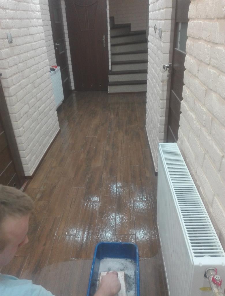 Czyszczenie podłogi z paneli imitujących drewno w korytarzu, widoczny grzejnik i fragment schodów, osoba myjąca podłogę z użyciem wiadra i gąbki.