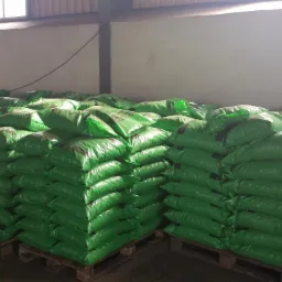 Magazyn: stosy zielonych worków na paletach, prawdopodobnie z zawartością do użyźniania gleby, w hali produkcyjnej, z widocznym oświetleniem.