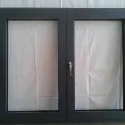 Dwuskrzydłowe okno w grafitowej ramie z widocznymi zawiasami i klamką, zabezpieczone folią ochronną.