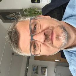 Portret mężczyzny w okularach na tle biura z obrazem budynku i rośliną doniczkową.