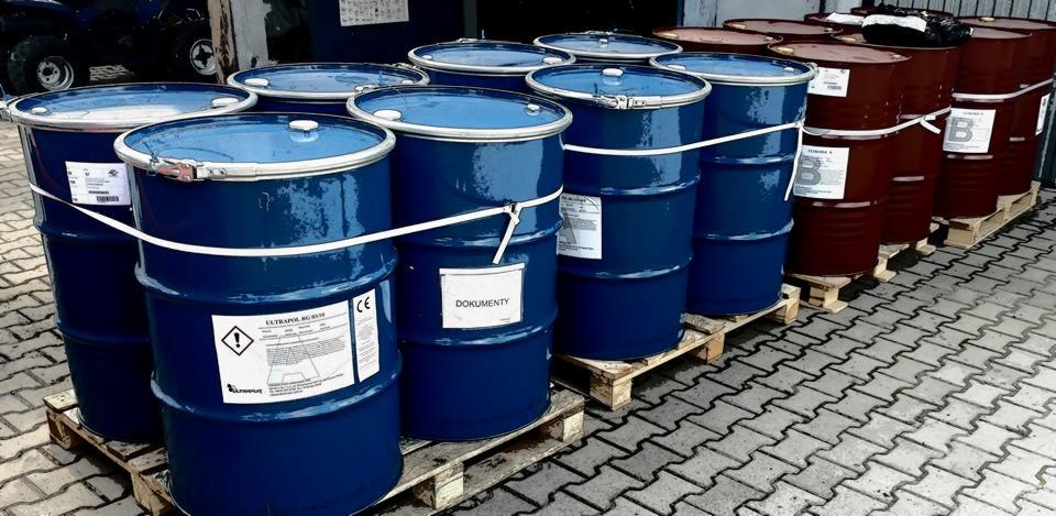 Błękitne i brązowe beczki z pianką poliuretanową na drewnianych paletach, etykiety informacyjne i ostrzegawcze na beczkach.