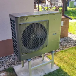 Zewnętrzna jednostka pompy ciepła NIBE AM S10 zamontowana na metalowej konstrukcji na betonowej podstawie, obok ściany budynku z małym otworem wentylacyjnym, na tle trawnika i drzew.