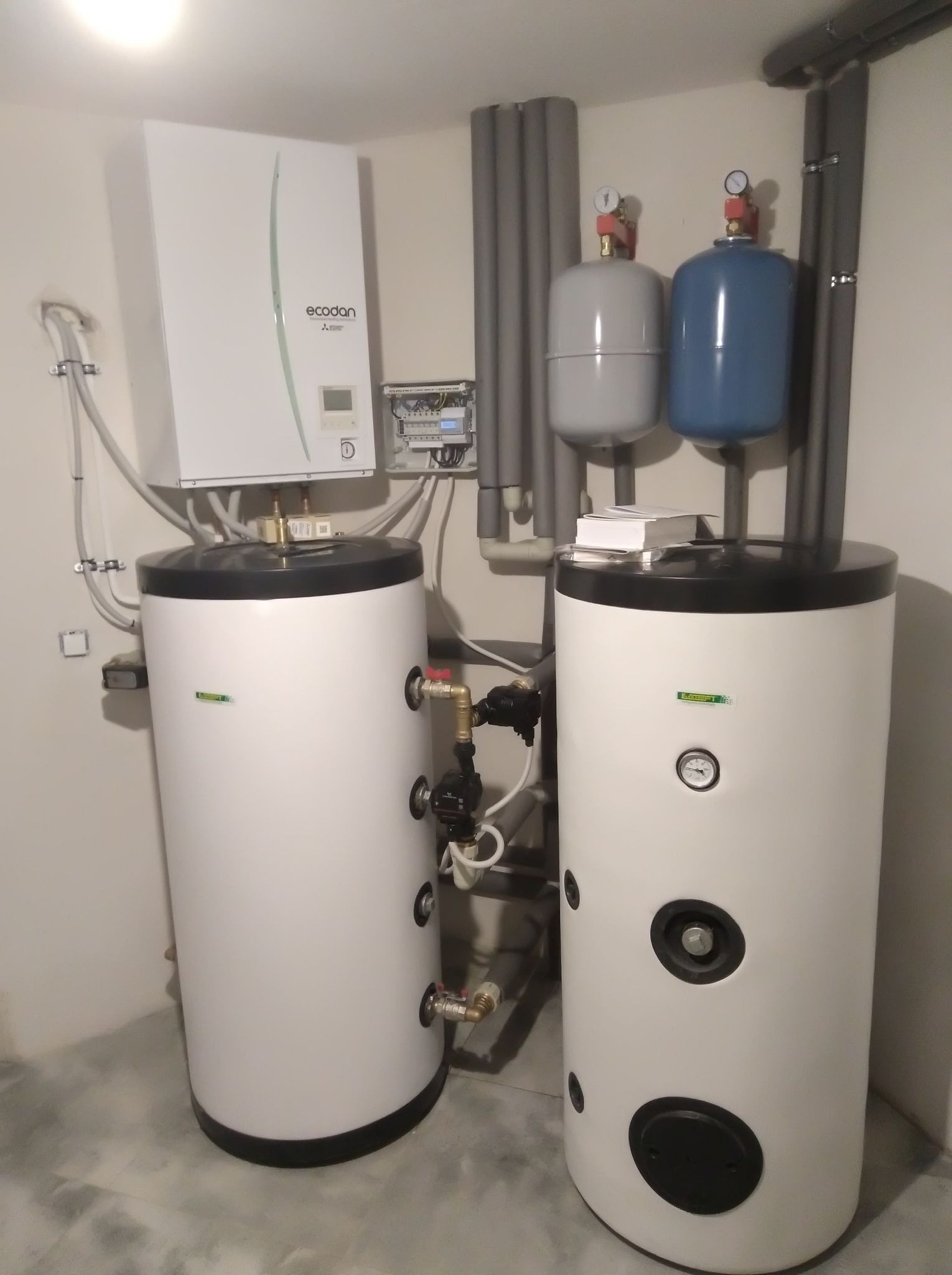 Dwa białe zbiorniki akumulacyjne z czarnymi pokrywami obok pompy ciepła marki Ecodan Mitsubishi w kotłowni, z widocznymi rurami instalacji grzewczej oraz zbiornikami wyrównawczymi.