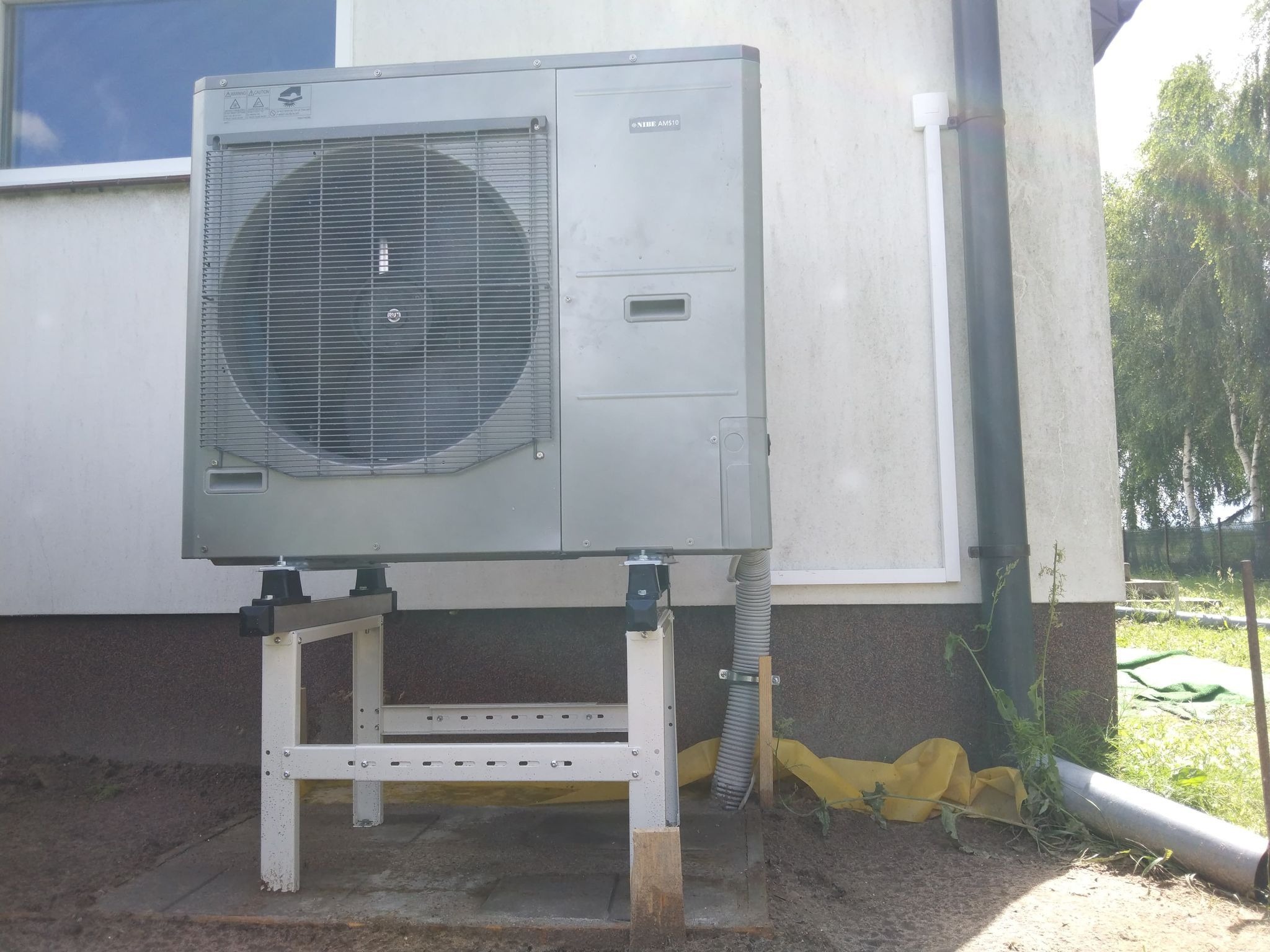 Szara jednostka zewnętrzna klimatyzacji Nibe AMS10 zamontowana na białym, metalowym stelażu przy ścianie budynku z widoczną rurą spustową i fragmentem okna, z zewnątrz zabezpieczona żółtą folią...