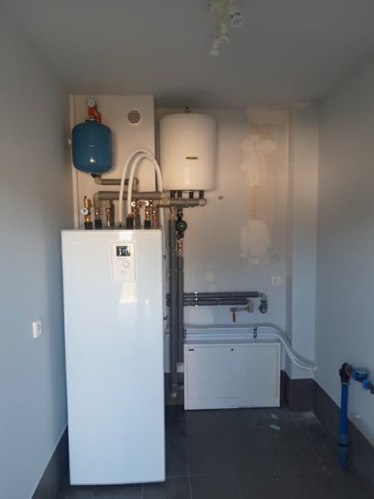 Biały piec centralnego ogrzewania z podłączonymi rurami i zbiornikiem wyrównawczym w pomieszczeniu z szarą podłogą i ścianami, widoczne ślady po malowaniu na ścianie.