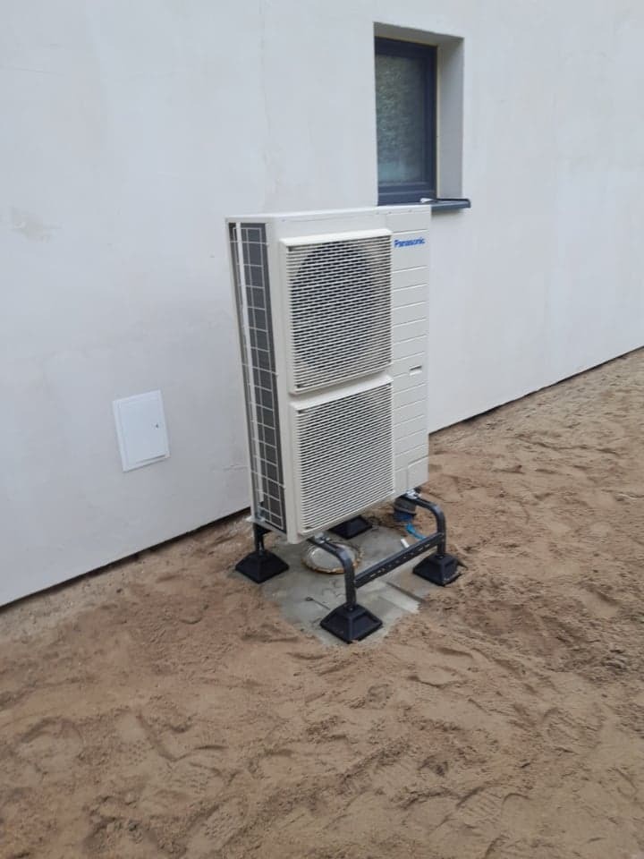 Biała jednostka zewnętrzna klimatyzacji Panasonic zamontowana na metalowej konstrukcji z czarnymi podstawami, stoi przy białej ścianie budynku z małym oknem, teren wokół wysypany piaskiem.