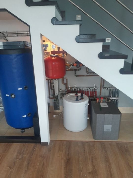 Instalacja grzewcza z zbiornikami i rozdzielaczem, umieszczona pod schodami w pomieszczeniu z panelami podłogowymi.