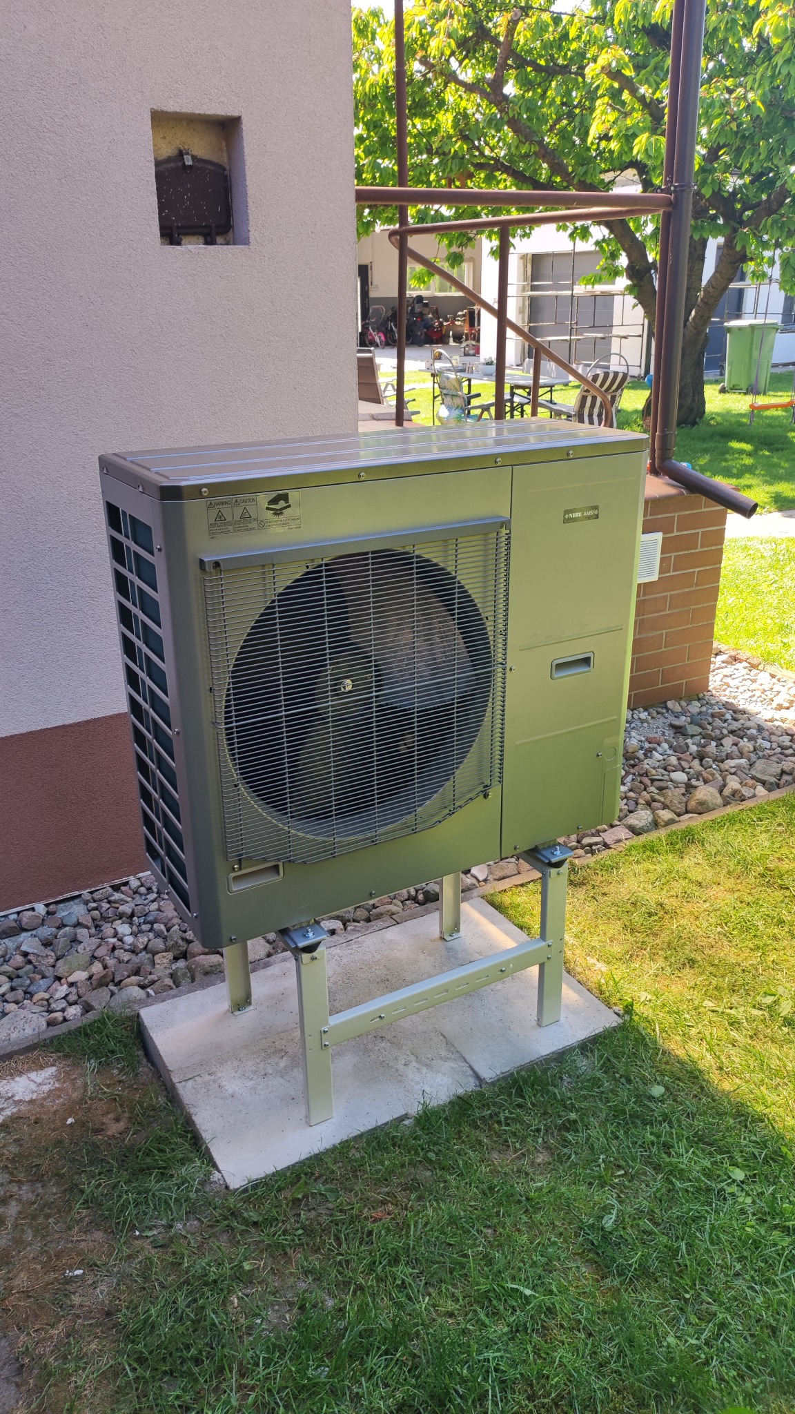 Zewnętrzna jednostka pompy ciepła NIBE AM S10 zamontowana na metalowej konstrukcji na betonowej podstawie, obok ściany budynku z małym otworem wentylacyjnym, na tle trawnika i drzew.