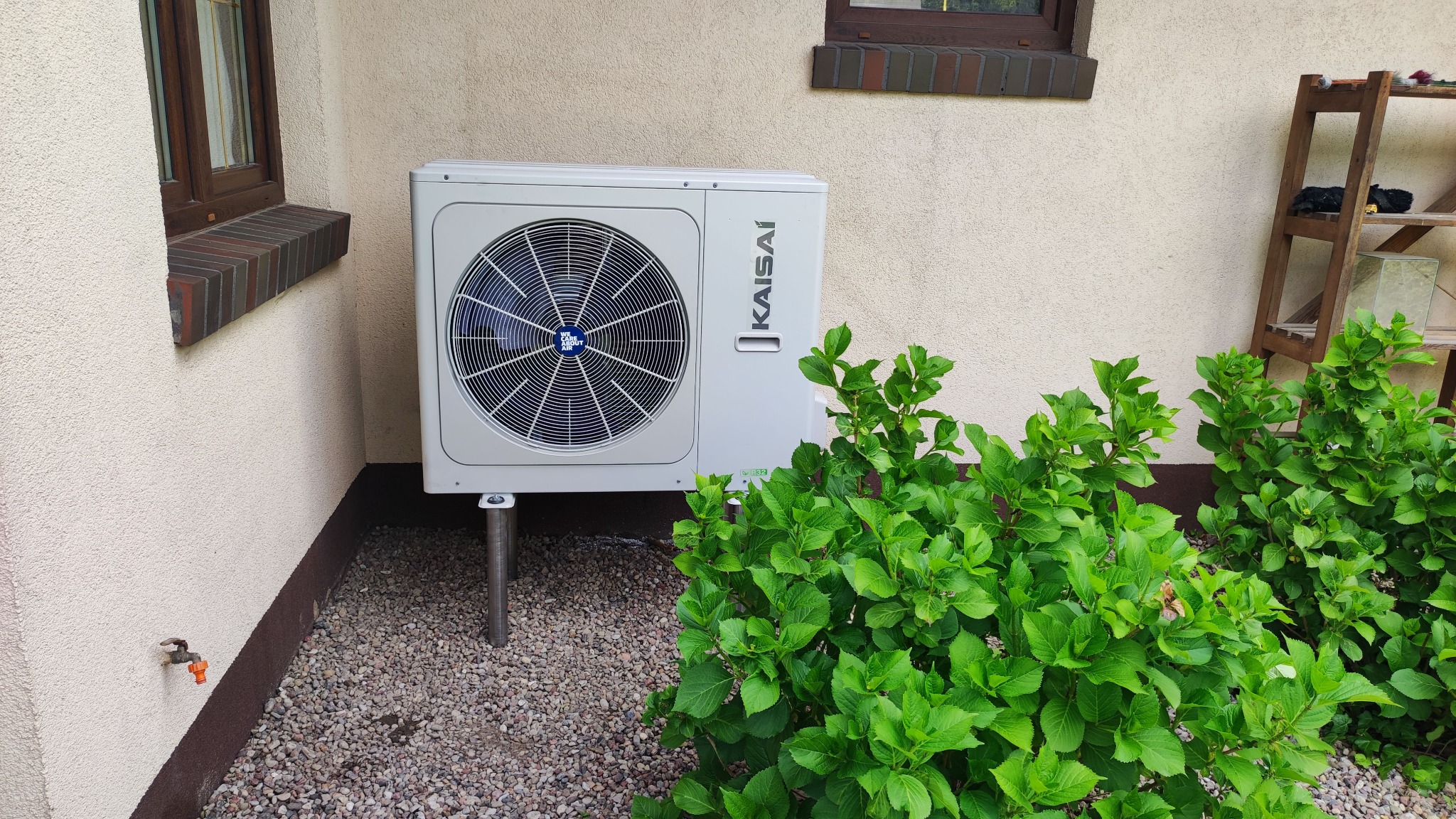 Biała jednostka zewnętrzna klimatyzacji Kaisai, zamontowana na metalowych nóżkach na żwirowym podłożu, obok ściany budynku z oknem i zielonymi krzewami hortensji.