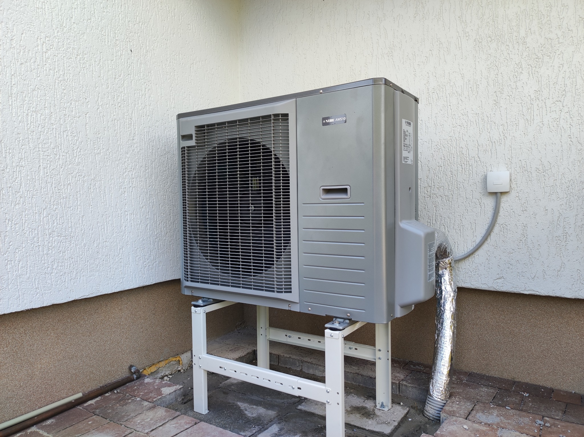 Zewnętrzna jednostka klimatyzacji Nibe AMS10 zamontowana na białym, metalowym stelażu przy ścianie budynku z fakturą baranka, widoczny przewód w srebrnej izolacji.