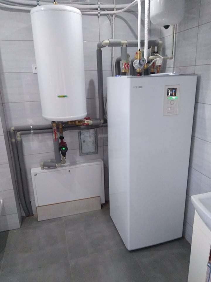 Biały piec centralnego ogrzewania z podłączonym zasobnikiem ciepłej wody, widoczne rury w izolacji, całość zamontowana w pomieszczeniu z szarymi płytkami.