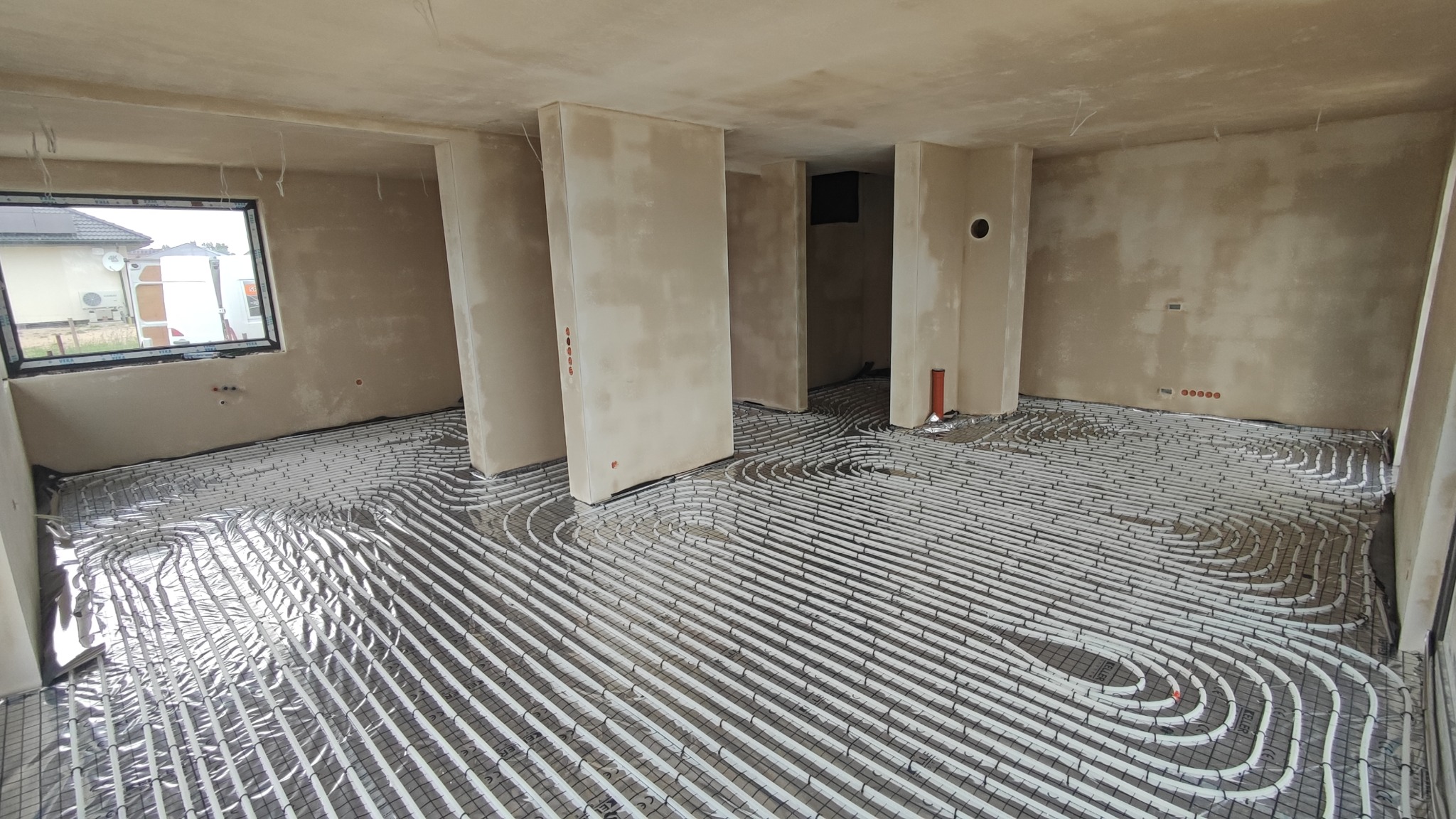 Rozłożona instalacja ogrzewania podłogowego na folii izolacyjnej w surowym stanie, widoczne rury w kształcie meandrów i fragment okna z widokiem na dom.