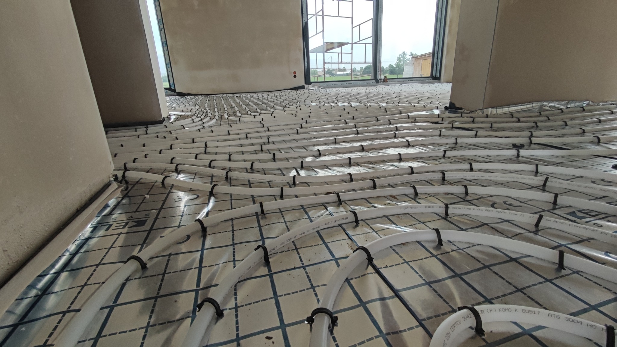 Białe rury ogrzewania podłogowego ułożone na folii z nadrukiem kratki, mocowane czarnymi klipsami, widok z niskiej perspektywy w surowym wnętrzu domu w budowie.