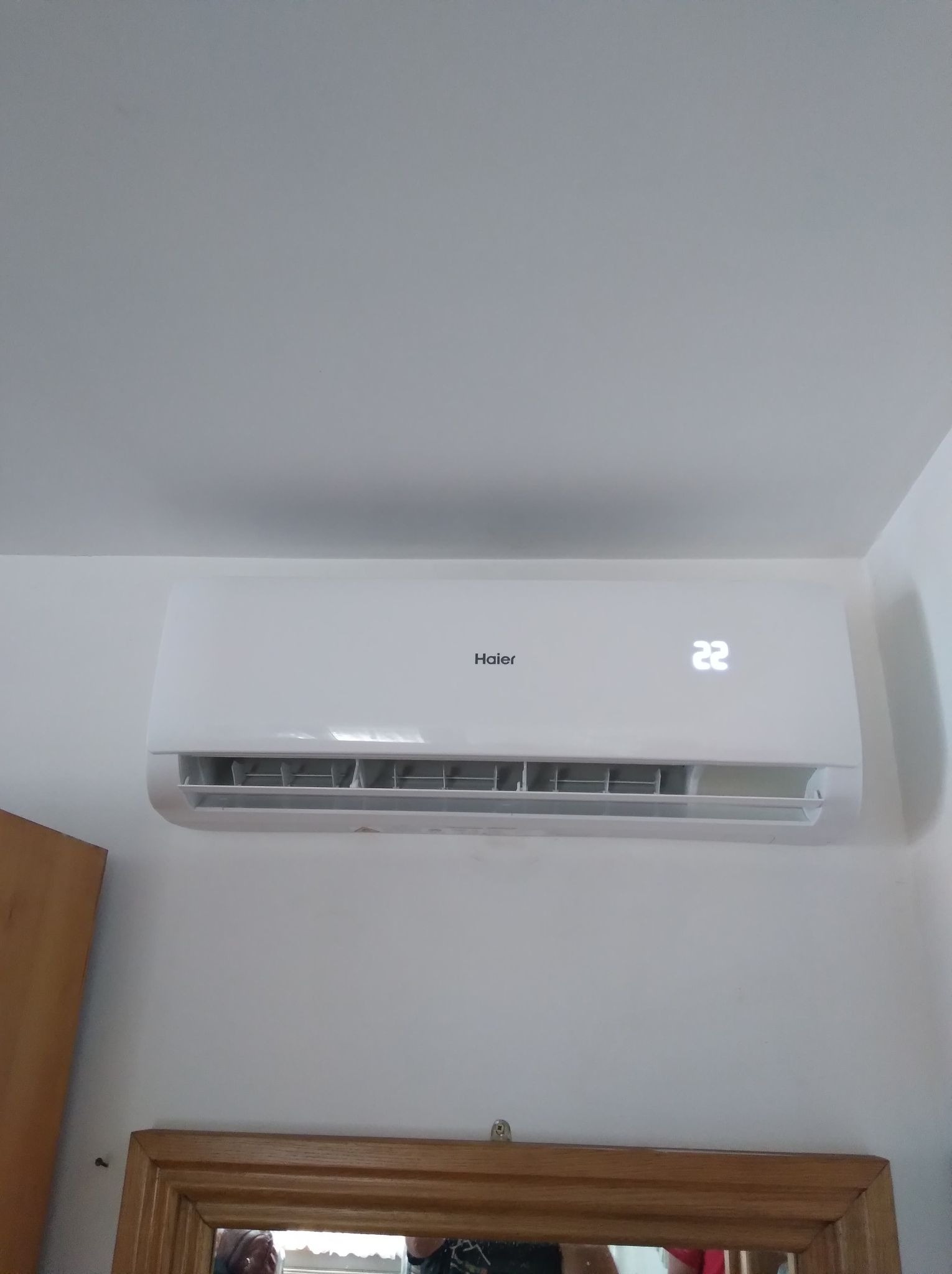 Biała klimatyzacja Haier zamontowana na białej ścianie, wyświetlająca temperaturę 22 stopnie, widoczne odbicie ramy lustra w dolnej części kadru.