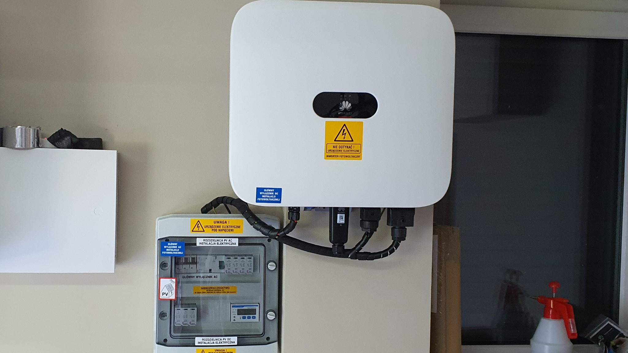 Biały inwerter fotowoltaiczny Huawei zamontowany obok rozdzielnicy PV AC/DC z widocznymi naklejkami ostrzegawczymi o napięciu elektrycznym.