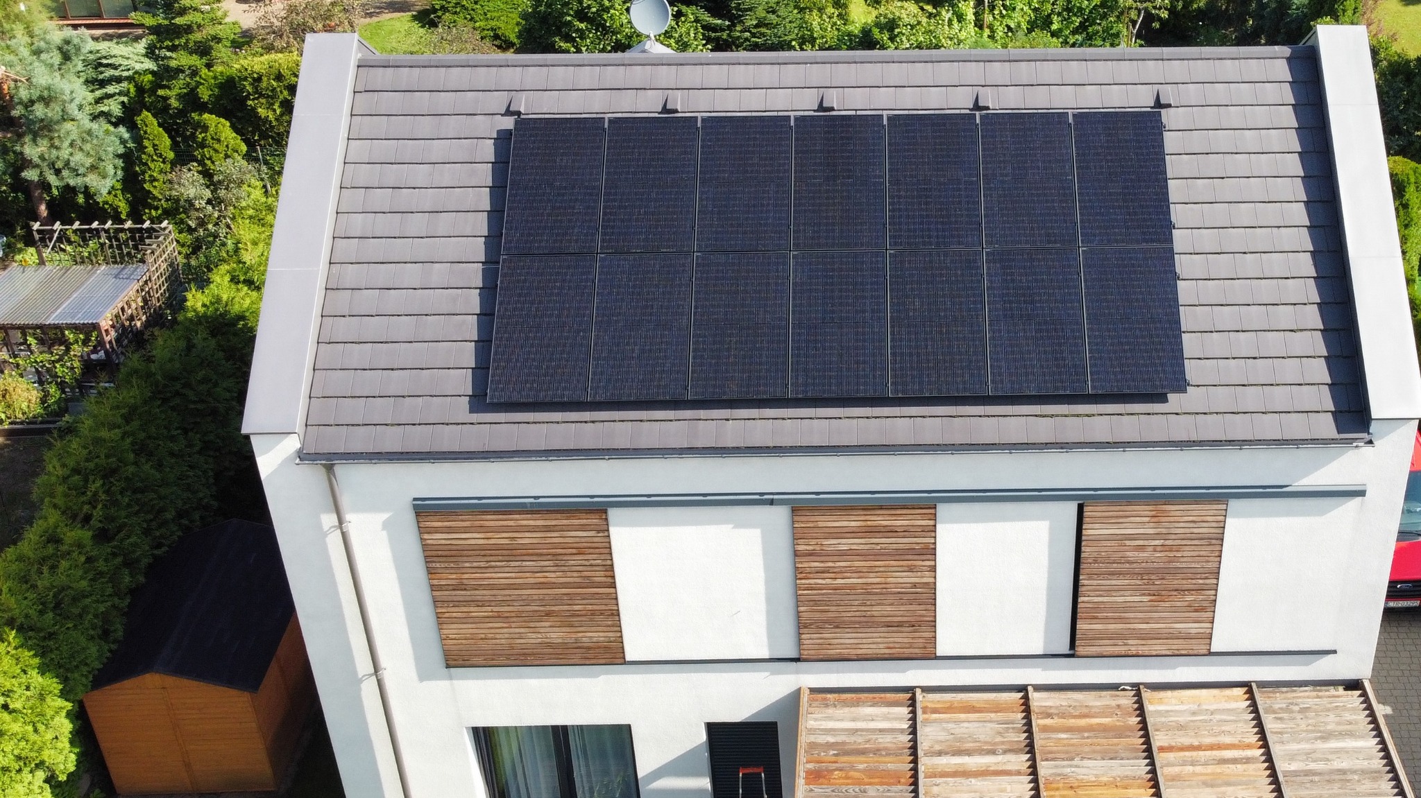 Widok z góry na dom z panelami słonecznymi na szarym dachu, drewniane elementy elewacji, antena satelitarna i zadbany ogród.